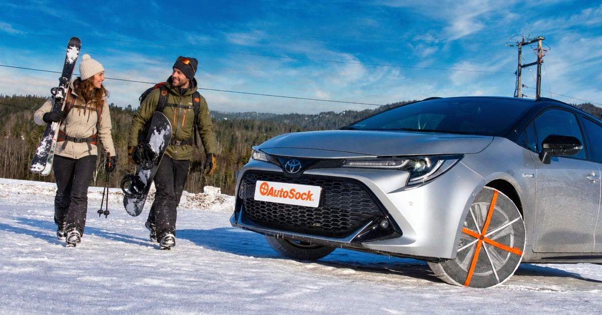 Toyota Corolla Hybrid plateado estacionado en la nieve bajo un cielo azul, con dos personas caminando cerca con esquís. La rueda delantera lleva fundas para nieve con franjas naranjas.
