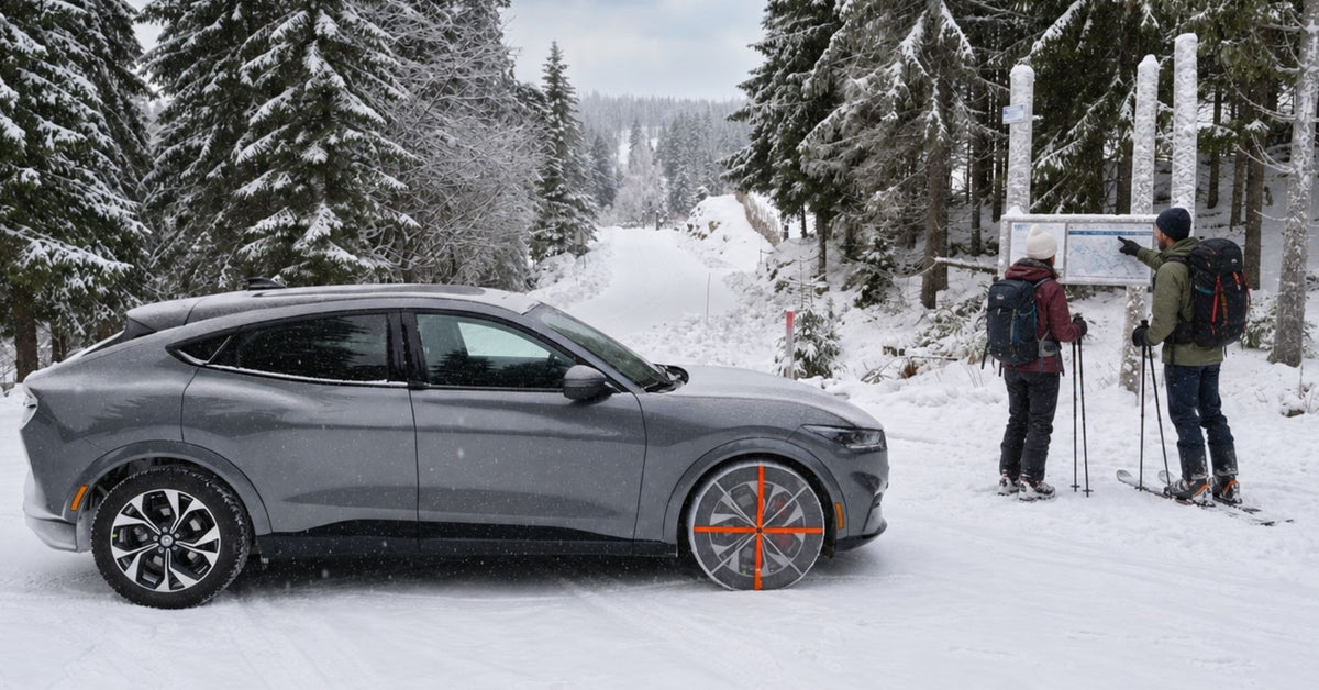 SUV Ford Mustang gris con cadenas de nieve textiles montadas, estacionado en una carretera de montaña nevada, con esquiadores cerca y rodeado de pinos cubiertos de nieve.