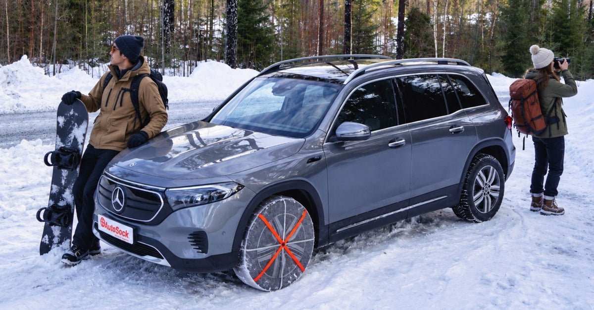 SUV Mercedes-Benz EQB gris estacionado en un entorno nevado con dos personas a su lado, una con una tabla de snowboard y la otra tomando una foto. El vehículo lleva fundas para nieve con tiras naranjas cruzadas.
