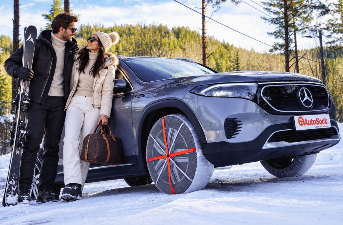 SUV eléctrico Mercedes-Benz gris con fundas para nieve instaladas en los neumáticos de invierno, estacionado en un entorno nevado con dos personas a su lado, una con esquís y la otra con un bolso marrón.